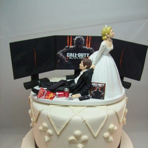 Puede incluir: Un pastel de boda de dos niveles decorado con glaseado blanco y una cinta negra. El nivel superior presenta una figura de novia y novio, el novio está acostado en un sofá con un control remoto, y la novia lo está tirando hacia arriba. El pastel está decorado con un tema de videojuego Call of Duty Black Ops.