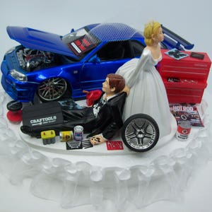 May include: Wedding cake topper featuring a blue sports car with the hood open, a bride, and a groom in a tuxedo. The groom is lying down with tools and a tire nearby. A red toolbox and a magazine are also present.
