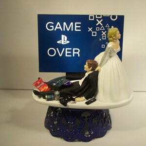 May include: A wedding cake topper featuring a bride pulling a groom away from a video game console. The groom is lying on a couch with a controller in his hand. The bride is wearing a white wedding dress and the groom is wearing a black tuxedo. The background is a blue screen with the words "GAME OVER" and a PlayStation logo.