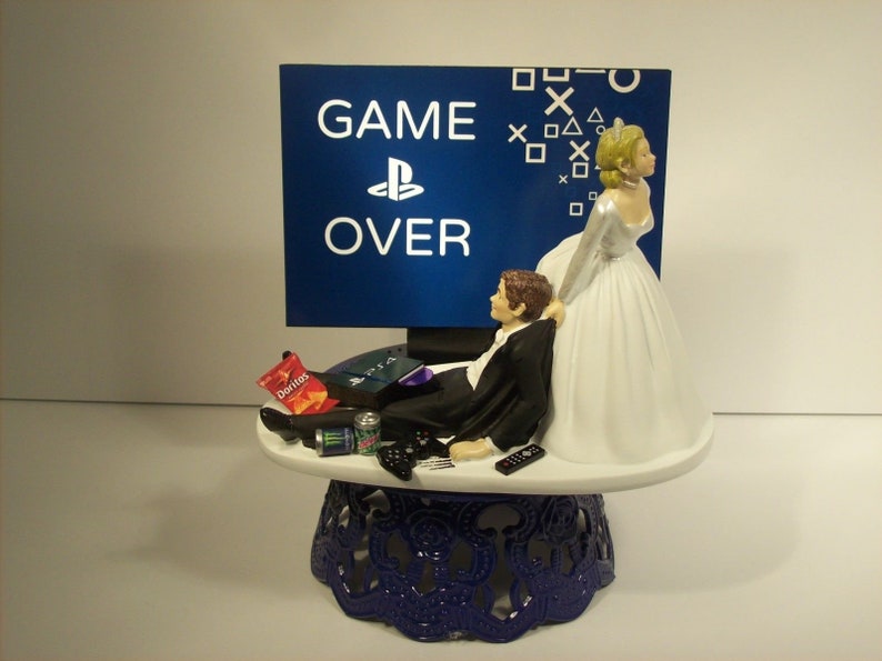 May include: A wedding cake topper featuring a bride pulling a groom away from a video game console. The groom is lying on a couch with a controller in his hand. The bride is wearing a white wedding dress and the groom is wearing a black tuxedo. The background is a blue screen with the words "GAME OVER" and a PlayStation logo.