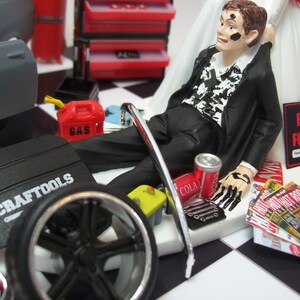 May include: A wedding cake topper featuring a groom dressed in a black suit with a white shirt and black tie. He is lying on his back with his hands up in the air. The cake topper also includes a car tire, a gas can, a toolbox, and a can of cola. The groom is covered in black paint and the cake topper has the words "Parts for Sale" on it.