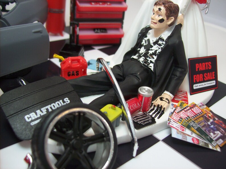 May include: A wedding cake topper featuring a groom dressed in a black suit with a white shirt and black tie. He is lying on his back with his hands up in the air. The cake topper also includes a car tire, a gas can, a toolbox, and a can of cola. The groom is covered in black paint and the cake topper has the words "Parts for Sale" on it.