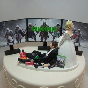 Puede incluir: Adorno para tarta de boda con novios. El novio está reclinado con aperitivos y un mando frente a tres pantallas con "Assassin's Creed" y "Game Over". La novia lo está tirando.