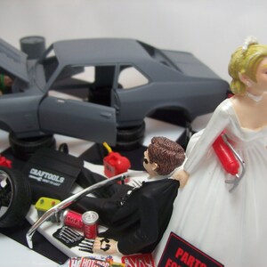 May include: A wedding cake topper featuring a bride and groom. The groom is dressed in a black tuxedo and is holding a wrench. The bride is wearing a white wedding dress and is holding a fire extinguisher. The cake topper also includes a gray Chevrolet Nova with the hood open, a toolbox, and other car parts.