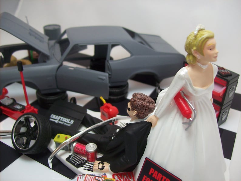 May include: A wedding cake topper featuring a bride and groom. The groom is dressed in a black tuxedo and is holding a wrench. The bride is wearing a white wedding dress and is holding a fire extinguisher. The cake topper also includes a gray Chevrolet Nova with the hood open, a toolbox, and other car parts.