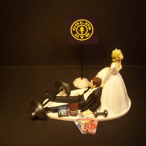 May include: A wedding cake topper featuring a bride and groom. The groom is lying on his back with a barbell over his chest. The bride is standing over him with her hand on his shoulder. The cake topper is decorated with a "Gold's Gym" logo and a magazine.