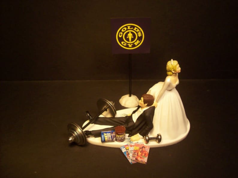 May include: A wedding cake topper featuring a bride and groom. The groom is lying on his back with a barbell over his chest. The bride is standing over him with her hand on his shoulder. The cake topper is decorated with a "Gold's Gym" logo and a magazine.