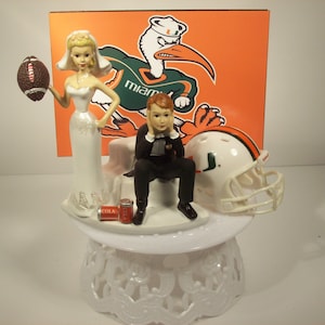 May include: A wedding cake topper featuring a bride and groom dressed in formal attire. The bride is holding a football and the groom is covering his ears. The background features a University of Miami logo and a football helmet.