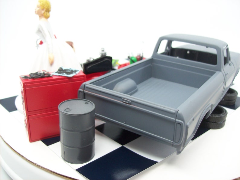 Może przedstawiać: Model ciężar&oacute;wki pick-up w kolorze szarym z czerwoną skrzynką na narzędzia, czarnym beczką na olej i białą figurką kobiety.  Ładowność ciężar&oacute;wki jest pusta, a ciężar&oacute;wka stoi na czarno-białej powierzchni w szachownicę.