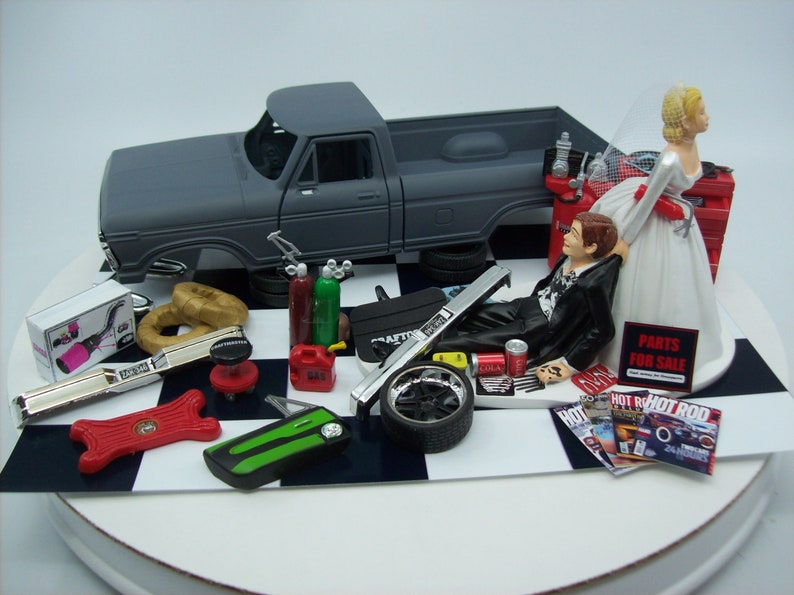 Może przedstawiać: Topper na tort weselny przedstawiający parę młodą z szarym pickupem, narzędziami i częściami samochodowymi. Pan młody leży na ziemi z puszką coli i czasopismem. Panna młoda stoi nad nim z tabliczką z napisem "Części na sprzedaż".