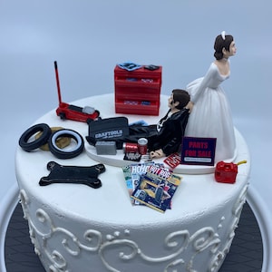 May include: A white wedding cake decorated with a bride and groom figurine. The groom is wearing a black suit and the bride is wearing a white dress. The cake is decorated with car parts, including tires, a jack, and a toolbox. The cake also has a sign that says "Parts for Sale".