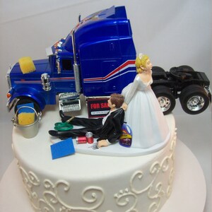 May include: A wedding cake topper featuring a bride and groom. The groom is lying on the ground next to a blue semi-truck, while the bride is standing and holding the truck. The cake is white with a decorative design.