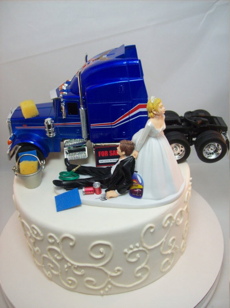 May include: A wedding cake topper featuring a bride and groom. The groom is lying on the ground next to a blue semi-truck, while the bride is standing and holding the truck. The cake is white with a decorative design.