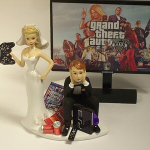 Puede incluir: Un adorno para pastel de boda con una novia sosteniendo un mando de videojuego y un novio sentado con una consola de videojuego y un mando. El fondo presenta una pantalla de televisión que muestra el videojuego "Grand Theft Auto V".