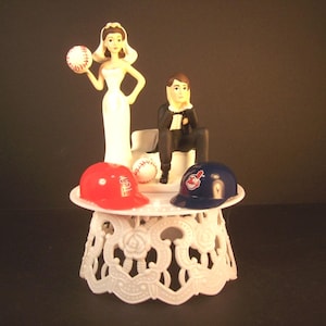 May include: A wedding cake topper featuring a bride in a white dress holding a baseball and a groom in a black tuxedo sitting on a bench. The figures are placed on a white decorative stand with a red baseball helmet and a blue baseball helmet.