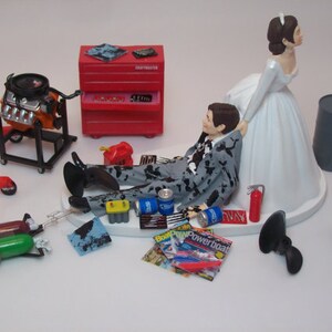 May include: A wedding cake topper featuring a bride and groom. The groom is lying on his back with a tool-covered suit, while the bride is standing over him with a hand on his shoulder. The cake topper includes a red toolbox, a car engine, and other tools.