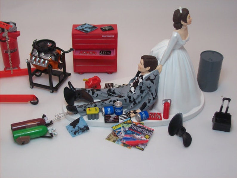 May include: A wedding cake topper featuring a bride and groom. The groom is lying on his back with a tool-covered suit, while the bride is standing over him with a hand on his shoulder. The cake topper includes a red toolbox, a car engine, and other tools.