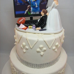 May include: A two-tiered white wedding cake with a silver ribbon around the bottom tier. The top tier features a bride and groom figurine. The groom is lying on a couch watching a football game on a television. The bride is pulling the groom away from the television. The cake is decorated with white frosting and a silver ribbon.