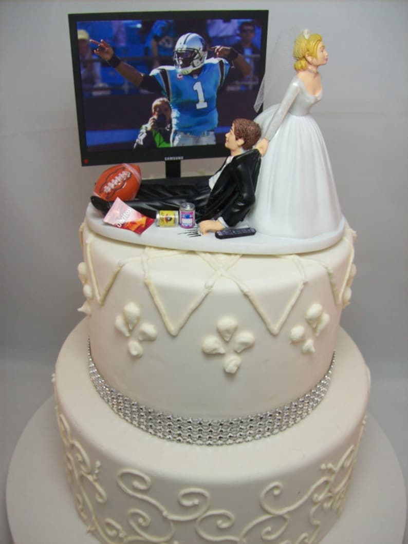 May include: A two-tiered white wedding cake with a silver ribbon around the bottom tier. The top tier features a bride and groom figurine. The groom is lying on a couch watching a football game on a television. The bride is pulling the groom away from the television. The cake is decorated with white frosting and a silver ribbon.