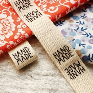 Puede incluir: Primer plano de etiquetas de tela color crema con las palabras "HAND MADE" impresas en negro. Las etiquetas están colocadas sobre telas estampadas en tonos rojos, blancos y azules. Las telas tienen diseños florales.