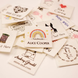Puede incluir: Una colección de pequeñas etiquetas de tela blanquecina con varios diseños y texto. Algunas etiquetas presentan ilustraciones como un arcoíris, una máquina de coser y un corazón. El texto incluye "handmade", "Alice Cooper" y "Audrey Greald."