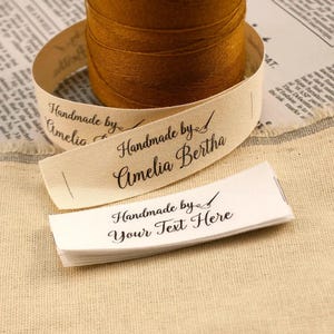 Peut inclure: Une bobine de fil marron avec un ruban crème portant l'inscription "Handmade by Amelia Bertha". Une pile d'étiquettes blanches avec le texte "Handmade by Your Text Here" est également visible. Les articles sont sur un tissu beige.