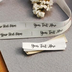 Peut inclure: Rubans et étiquettes en tissu blanc avec le texte "Your Text Here" imprimé en script noir. Le ruban est enroulé et les étiquettes sont empilées. Quelques fleurs blanches sont en arrière-plan.