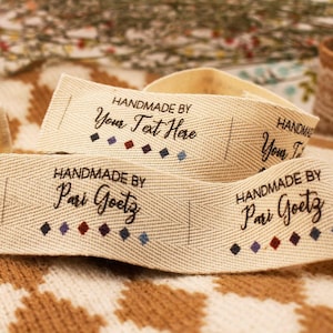 Könnte beinhalten: Nahaufnahme von beigen Stoffetiketten mit dem Text "HANDMADE BY Your Text Here" und "HANDMADE BY Pari Goetz" in schwarzer Schrift. Jedes Etikett hat eine Reihe kleiner, farbiger Diamanten.