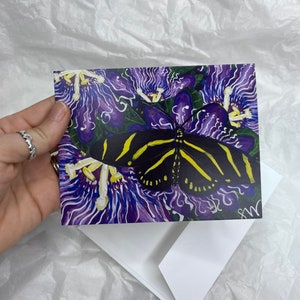 May include: A black and yellow butterfly with white stripes on its wings is perched on a purple flower with white accents. The flower has a swirling pattern and is surrounded by green leaves.