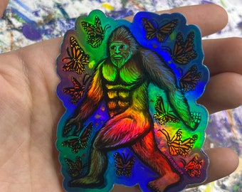 May include: Holographic sticker featuring a colorful Bigfoot surrounded by monarch butterflies. The artwork has a rainbow gradient effect, with the Bigfoot in shades of orange, yellow, and green. The butterflies are orange and black. The sticker has a glossy finish.