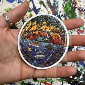 May include: A round sticker featuring a coastal scene with a pelican, fish, dolphins, and a turtle. The artwork is framed by trees and a sunset sky. The sticker has a white border and is held in a hand.