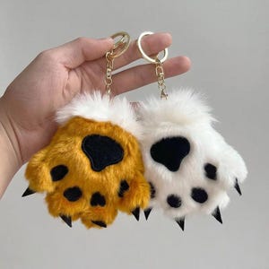 May include: Two plush paw keychains, one golden yellow and one white, each with black paw pads and claws. The keychains have gold-colored metal rings for attaching to keys or bags. The paws have a fluffy texture.