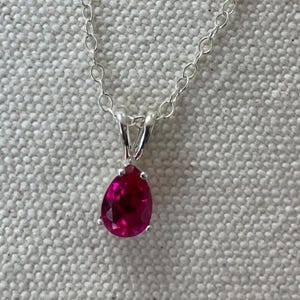 May include: A silver necklace with a pear-shaped, vibrant red gemstone pendant. The pendant is set in a silver bezel and hangs from a delicate silver chain. The necklace is displayed on a neutral-colored background.