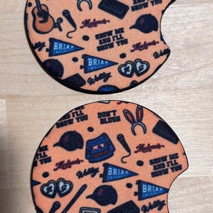 May include: Two circular car cup holder coasters featuring a peach-coloured background with a repeating pattern of blue jerseys with the number 44, acoustic guitars, microphones, bunny ears, baseball caps, pennants labelled BRIAN, and text reading SHOW ME AND I'LL SHOW YOU.