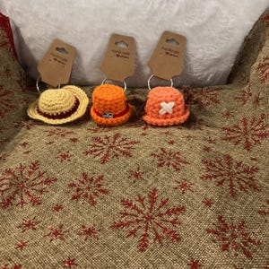 Puede incluir: Tres llaveros de sombrero de ganchillo hechos a mano en amarillo, naranja y coral. Cada sombrero tiene un diseño único, incluyendo una cruz, ojos y un ala clásica. Los llaveros están unidos a etiquetas marrones que dicen "Handmade with love."