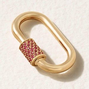 Könnte beinhalten: Goldfarbener, oval geformter Schmuckverschluss mit einem zylindrischen Abschnitt, der mit kleinen, roten Edelsteinen besetzt ist. Der Verschluss ist vor einem weißen Hintergrund platziert, was seine polierte Oberfläche und sein aufwendiges Design hervorhebt.