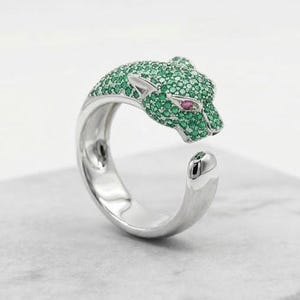 Könnte beinhalten: Ein silberner Ring mit einem Pantherkopf-Design. Der Pantherkopf ist mit grünen und rosa Edelsteinen besetzt. Der Ring wird auf einem Marmorblock präsentiert.