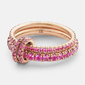 Könnte beinhalten: Ein Ring-Set aus Roségold mit drei Bändern, von denen zwei mit kleinen, leuchtend rosa Edelsteinen besetzt sind. Die Bänder sind miteinander verflochten und bilden an einer Seite ein knotenartiges Design. Der Ring wird vor einem schlichten Hintergrund präsentiert.