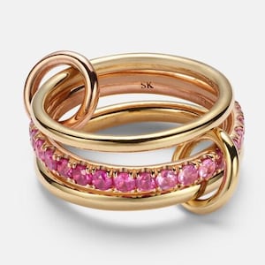 Könnte beinhalten: Ein goldener Ringsatz mit drei ineinandergreifenden Bändern. Ein Band ist mit kleinen rosa Edelsteinen besetzt. Die Ringe sind durch kleine goldene Schlaufen verbunden. Die Ringe sind ein Schmuckstück.