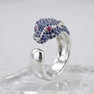 Könnte beinhalten: Ein silberner Ring mit einem Pantherkopf-Design. Der Pantherkopf ist mit blauen und roten Edelsteinen verziert. Der Ring wird auf einer klaren, strukturierten Oberfläche präsentiert, mit einem kleinen Pfotenabdruck-Anhänger in der Nähe.