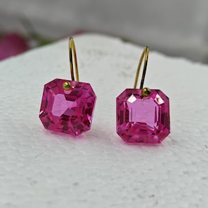 Könnte beinhalten: Ein Paar rosa Edelstein-Ohrringe mit goldfarbenen Haken. Jeder Ohrring hat einen facettierten, rechteckigen rosa Stein. Die Ohrringe sind vor einem weißen Hintergrund platziert, der die Farbe und das Design hervorhebt.