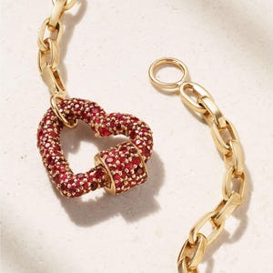 Könnte beinhalten: Goldfarbenes Kettenarmband mit herzförmigem Verschluss. Das Herz ist mit kleinen, roten Edelsteinen besetzt. Die Kettenglieder sind oval und der Verschluss ist offen.