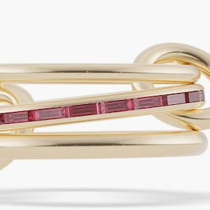 Op de afbeelding: Een goudkleurige ring met een uniek in elkaar grijpend ontwerp. De ring heeft een centrale balk versierd met rechthoekige, roze edelstenen. De algehele esthetiek is modern en elegant, geschikt voor diverse gelegenheden.