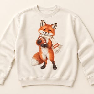 Peut inclure: Un sweat-shirt à col rond couleur crème arborant une illustration centrale d'un renard roux à la queue blanche. Le renard porte une écharpe couleur crème à rayures orange et tient un ballon de football américain marron entre ses pattes.