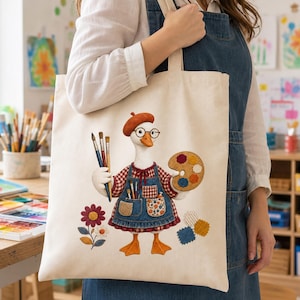 Peut inclure: Un sac fourre-tout en toile couleur crème orné d'une broderie représentant une oie blanche portant un béret rouge, des lunettes et un tablier en jean. L'oie tient trois pinceaux et une palette en bois, entourée d'une fleur et d'échantillons.