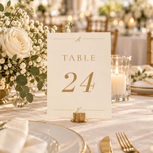 Könnte beinhalten: Eine rechteckige, cremefarbene Tischnummernkarte mit dem Text "TABLE 24" in goldener Schrift. Die Karte wird von einem kleinen, runden Ständer aus goldfarbenem Metall aufrecht gehalten und ist mit dünnen horizontalen Linien und kleinen Blumen verziert.