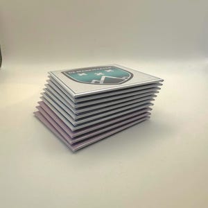 Op de afbeelding: Een stapel vierkante onderzetters met een teal, wit en zwart embleemdesign. Het embleem toont drie witte kruisen en de tekst "DE MARKIEZATEN". De onderzetters hebben een zilveren rand en een lichtpaarse onderkant.