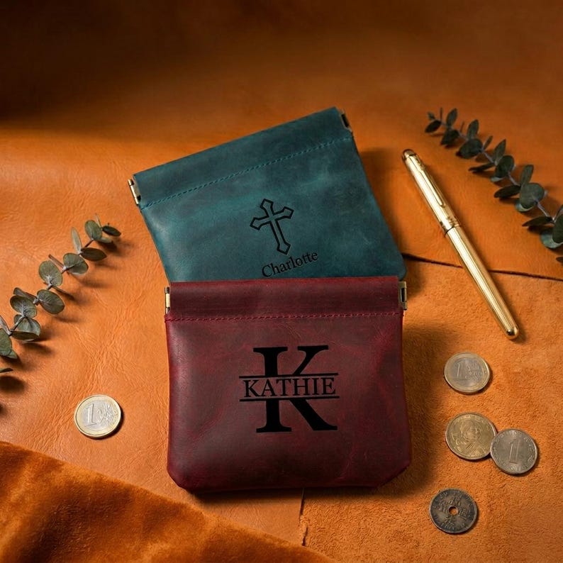 May include: Two leather coin purses, one teal and one burgundy, with gold-colored clasps. The teal purse has a cross and the name "Charlotte" engraved. The burgundy purse features the name "Kathie" with a large initial "K".