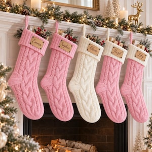 Op de afbeelding: Vijf kerstsokken met kabelbreisel hangend op een rij. Drie zijn roze en twee zijn wit. Elke sok is voorzien van een rechthoekig houten naamplaatje waarop de namen Emma, Sophia, Oliver, Hazel en Lily in het hout zijn gegraveerd.