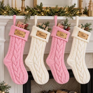 Op de afbeelding: Vier kerstsokken van kabelbreisel in afwisselende roze en crèmekleuren, elk voorzien van een geribbelde boord met een rechthoekig leren label waarop de namen Claire, Mason, Isla en Theo zijn gegraveerd, en kleine cadeautjes erin.